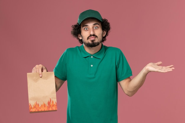 Vue de face de courrier masculin en uniforme vert et cape tenant un paquet de papier alimentaire et posant avec une expression confuse sur le travail de livraison uniforme de service de fond rose