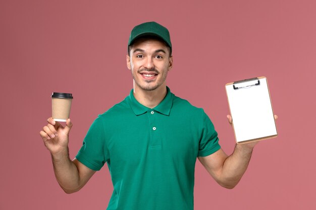 Vue de face de courrier masculin en uniforme vert tenant la tasse de café de livraison et le bloc-notes avec sourire sur le bureau rose