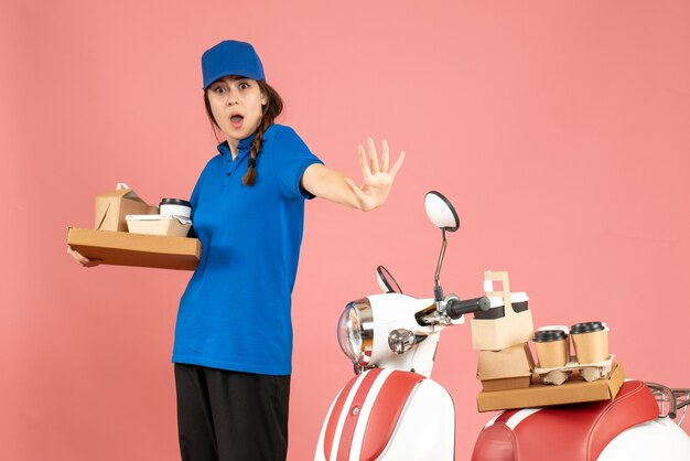 Vue de face d'une curieuse coursière debout à côté d'une moto tenant du café et de petits gâteaux sur fond de couleur pêche pastel