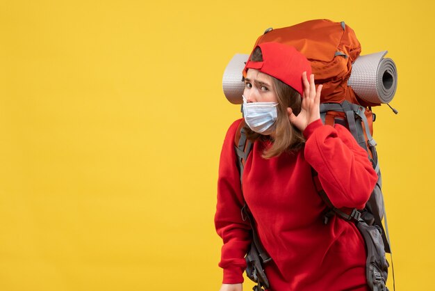 Vue de face curieuse jeune fille avec sac à dos touristique et masque en écoutant quelque chose