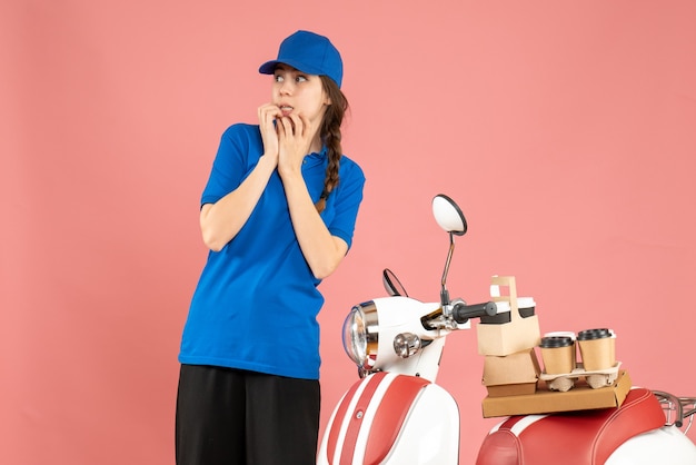 Vue De Face D'une Dame De Messagerie Confuse Debout à Côté D'une Moto Avec Du Café Et Des Petits Gâteaux Dessus Sur Fond De Couleur Pêche Pastel