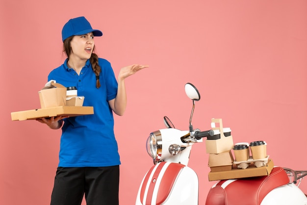 Vue de face d'une dame de messagerie confuse debout à côté d'une moto tenant du café et de petits gâteaux sur fond de couleur pêche pastel