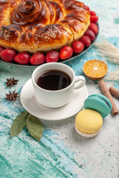 Vue de face délicieuse tarte sucrée avec des macarons aux fraises et tasse de thé sur plancher bleu