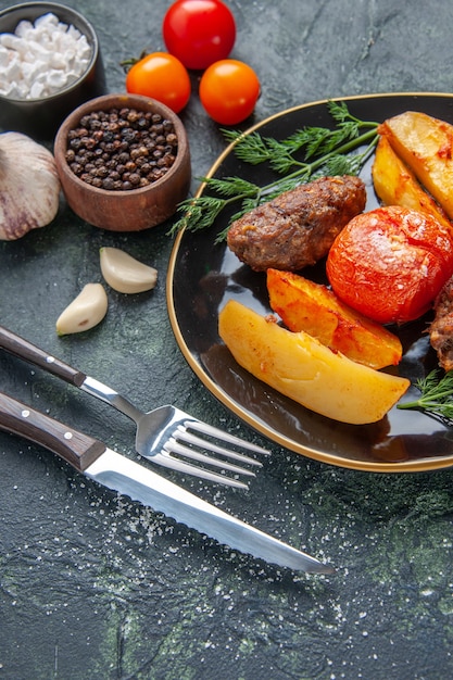 Vue de face de délicieuses escalopes de viande cuites au four avec des pommes de terre et des tomates sur une assiette noire ensemble de couverts épices ails tomates sur fond de couleurs mélangées vert noir