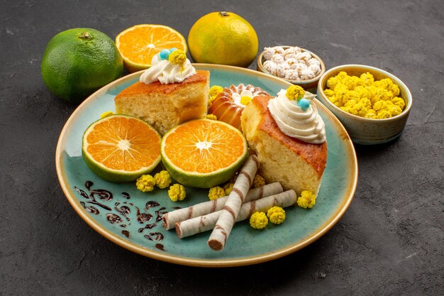 Vue de face de délicieuses tranches de tarte avec des bonbons et des mandarines fraîches sur l'espace sombre