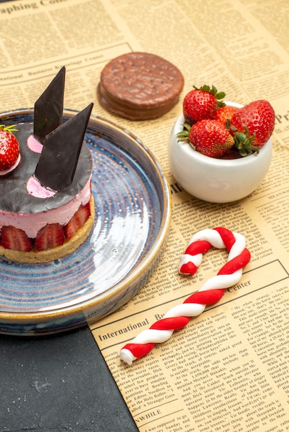Vue de face délicieux cheesecake à la fraise et au chocolat sur une assiette bol de fraises biscuit bonbons de Noël un journal sur fond sombre isolé
