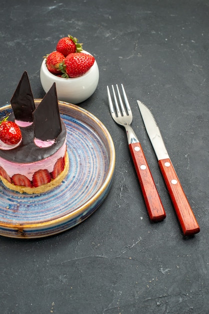 Vue de face délicieux cheesecake à la fraise et au chocolat sur un bol d'assiette avec un couteau à fourche à fraises sur l'obscurité