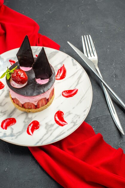 Vue de face délicieux cheesecake avec fraise et chocolat sur plaque châle rouge croisé couteau et fourchette sur fond sombre isolé