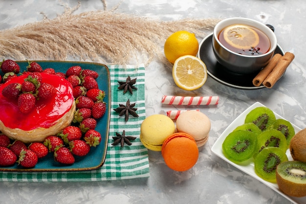 Vue de face délicieux gâteau aux fraises avec des fraises fraîches tasse de thé et macarons français sur bureau blanc