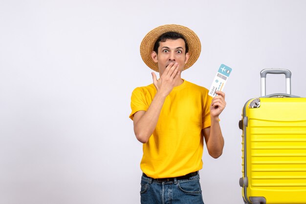 Vue de face demandé jeune homme en t-shirt jaune debout près de valise jaune tenant un billet