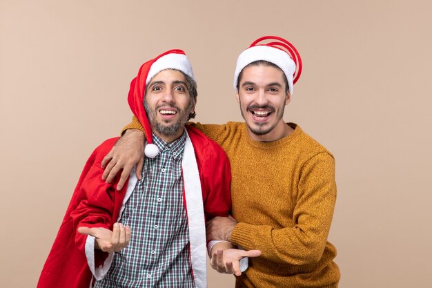 Vue de face deux amis avec des chapeaux de père Noël souriant sur fond isolé beige