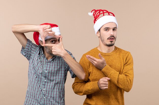 Vue de face deux gars avec des chapeaux de père Noël l'un faisant signe de l'appareil photo avec ses mains et l'autre lui montrant sur fond isolé beige