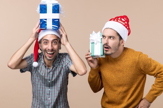 Vue de face deux gars avec des chapeaux de père Noël l'un mettant présent sur sa tête l'autre en pensant à ce sujet sur fond isolé beige