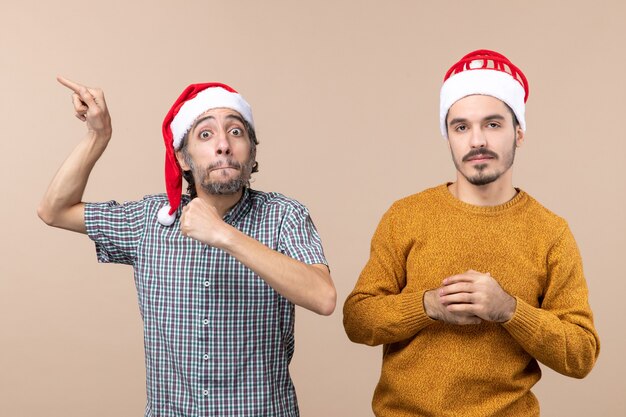 Vue de face deux gars avec des chapeaux de père Noël un montrant quelque chose avec un grand intérêt en se tenant debout sur fond isolé beige