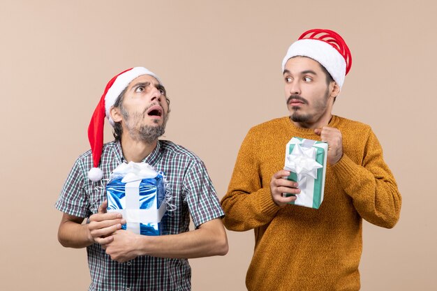 Vue de face deux gars confus portant des chapeaux de père Noël et tenant des cadeaux de Noël sur fond isolé beige