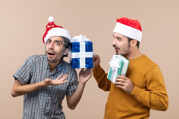Vue de face deux gars confus portant des chapeaux de père Noël et vérifier leurs cadeaux de Noël sur fond isolé beige