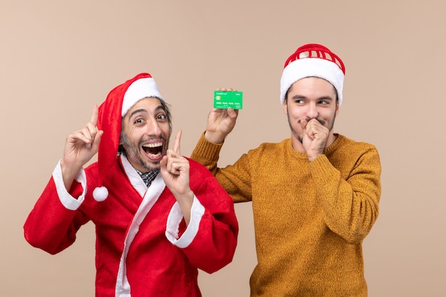 Vue de face deux gars heureux l'un avec manteau de père Noël et l'autre avec carte de crédit fermant la bouche sur fond isolé beige