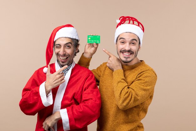 Vue de face deux gars heureux l'un avec manteau de père Noël et l'autre avec carte de crédit souriant sur fond isolé beige