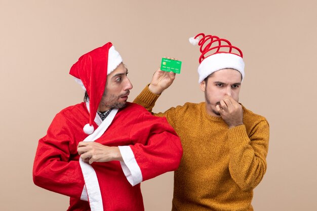 Vue de face deux gars de Noël un avec manteau de père Noël et l'autre avec carte de crédit fermant son nez sur fond isolé beige