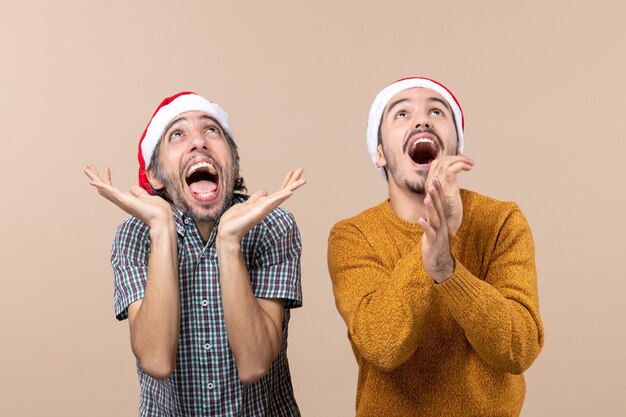 Vue de face deux mecs exaltés avec des chapeaux de père Noël applaudissant dans leurs mains en se tenant debout sur fond isolé beige