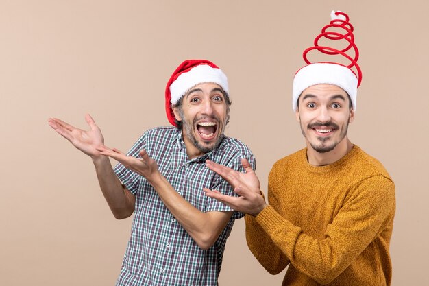 Vue de face deux mecs heureux avec des chapeaux de père Noël montrant quelque chose sur fond isolé beige