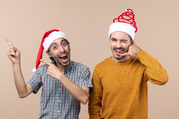 Vue de face deux mecs souriants avec des chapeaux de père Noël l'un montrant quelque chose et l'autre faisant appelez-moi téléphone signe sur fond isolé beige