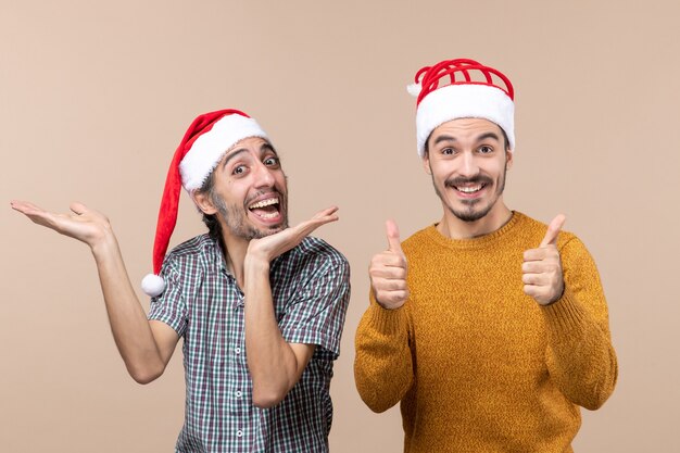 Vue de face deux mecs souriants avec des chapeaux de père Noël l'un montrant quelque chose de l'autre faisant signe avec les deux mains sur fond beige