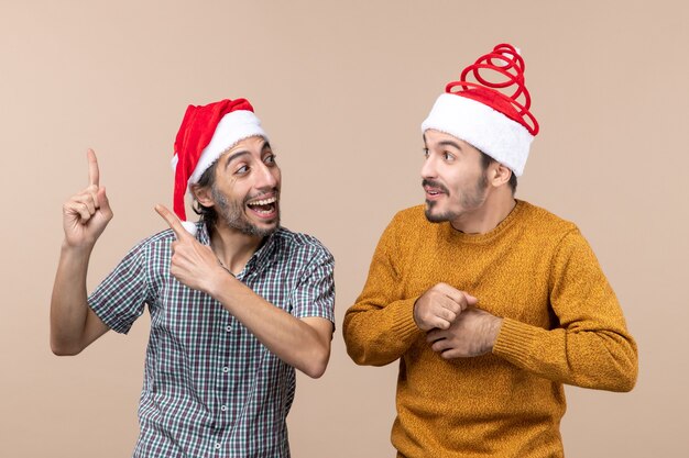 Vue de face deux mecs souriants avec des chapeaux de père Noël l'un montrant quelque chose à l'autre sur fond isolé beige