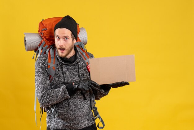 Vue de face du beau mâle auto-stoppeur avec des gants en cuir et sac à dos tenant un carton vierge