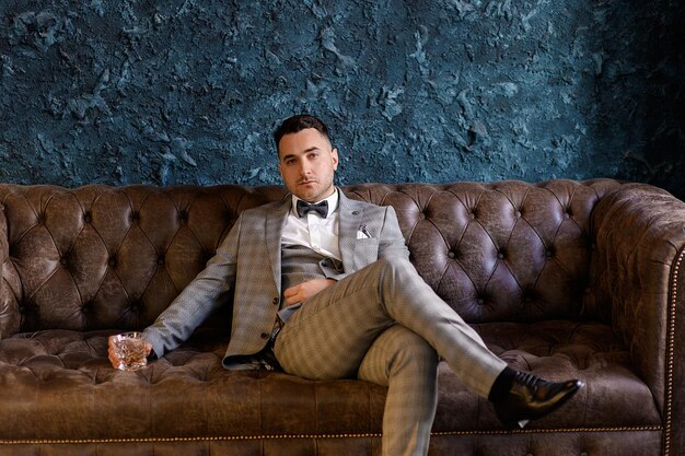 Vue de face du beau marié avec barbe portant un costume à la mode assis sur un canapé en cuir dans un appartement de luxe tenant un verre rempli de boisson et regardant la caméra