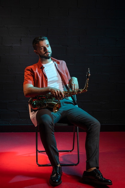 Vue de face du beau musicien assis sur une chaise tout en tenant le saxophone