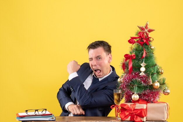 Vue de face du bon homme montrant le geste gagnant assis à la table près de l'arbre de Noël et présente sur jaune