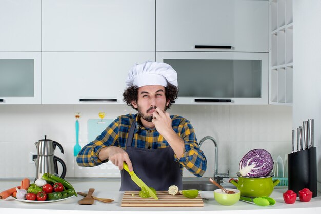 Vue de face du chef masculin au visage aigre avec des légumes frais et cuisine avec des ustensiles de cuisine et dégustation de poivrons verts dans la cuisine blanche