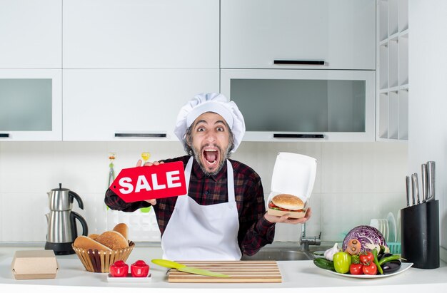 Vue de face du chef masculin hurlant brandissant une pancarte de vente et un hamburger dans la cuisine