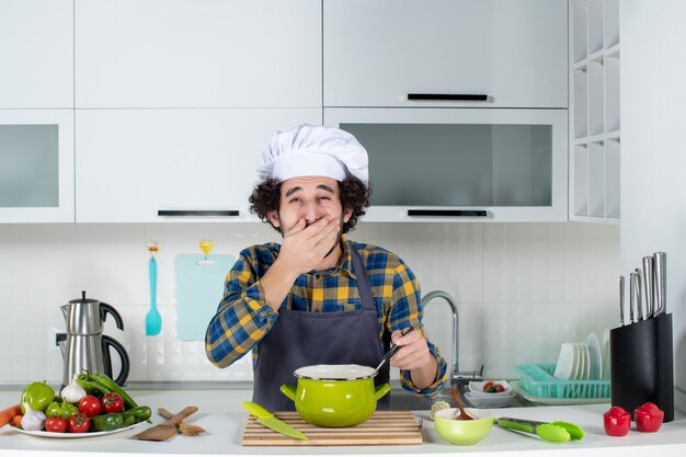 Vue de face du chef masculin avec des légumes frais et tenant une cuillère dans le repas se sentant fatigué dans la cuisine blanche