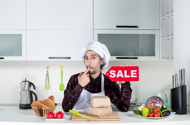 Vue de face du chef masculin sceptique en uniforme brandissant un panneau de vente rouge mettant le doigt dans sa bouche dans une cuisine moderne