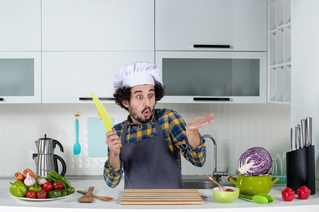 Vue de face du chef masculin se demandant avec des légumes frais et cuisinant avec des ustensiles de cuisine et tenant un couteau dans la cuisine blanche