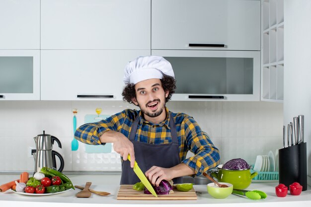 Vue de face du chef souriant avec des légumes frais coupant des aliments dans la cuisine blanche