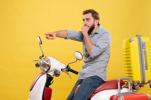 Vue de face du concept de voyage avec un jeune homme choqué assis sur une moto avec des valises pointant vers l'avant sur elle sur jaune