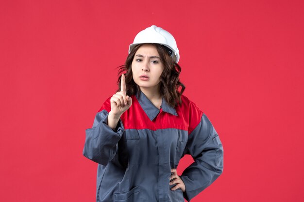 Vue de face du constructeur féminin nerveux en uniforme avec un casque et pointant vers le haut sur fond rouge isolé