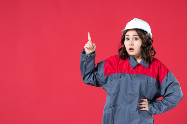 Vue de face du constructeur féminin surpris en uniforme avec un casque et pointant vers le haut sur fond rouge isolé