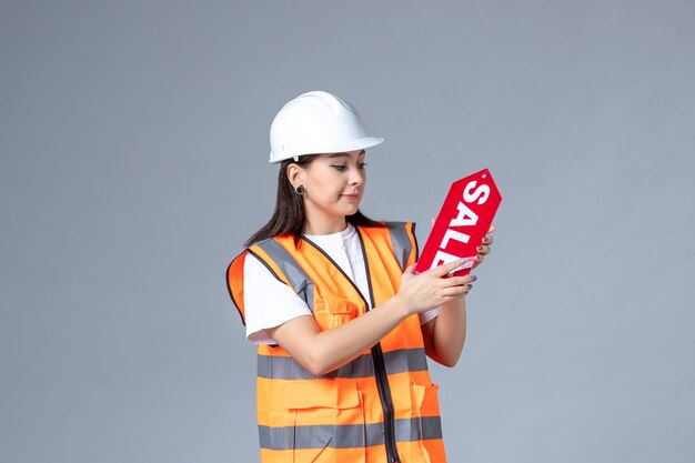 Vue de face du constructeur féminin tenant un tableau de vente rouge sur un mur gris