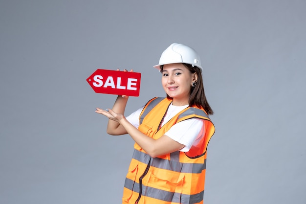 Vue de face du constructeur féminin tenant un tableau de vente rouge sur un mur gris