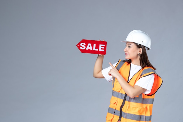 Vue de face du constructeur féminin tenant un tableau de vente rouge sur un mur gris