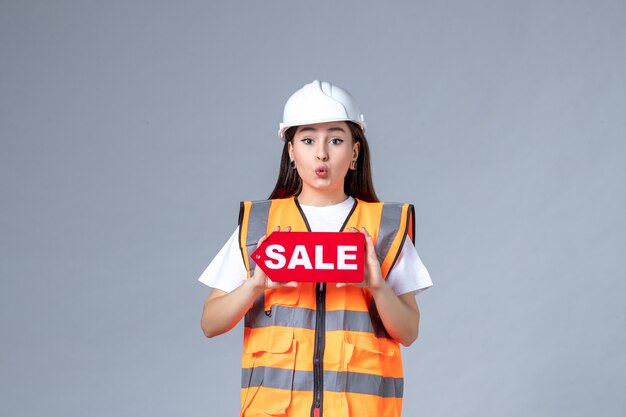 Vue de face du constructeur féminin tenant un tableau de vente rouge sur un mur gris