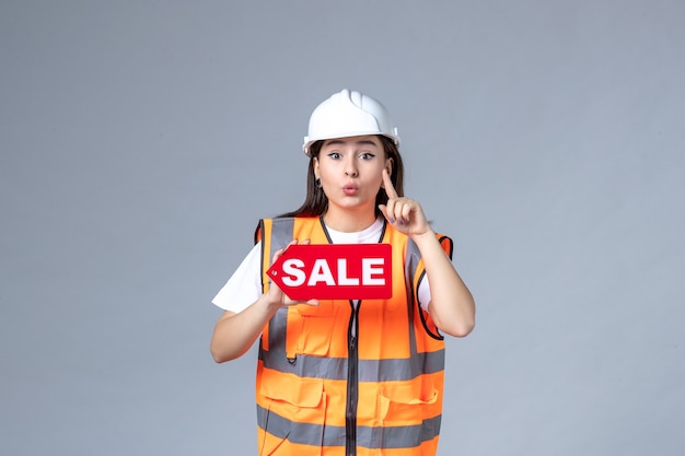 Vue de face du constructeur féminin tenant un tableau de vente rouge sur un mur gris