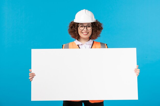 Vue de face du constructeur féminin en uniforme tenant un bureau blanc sur un mur bleu