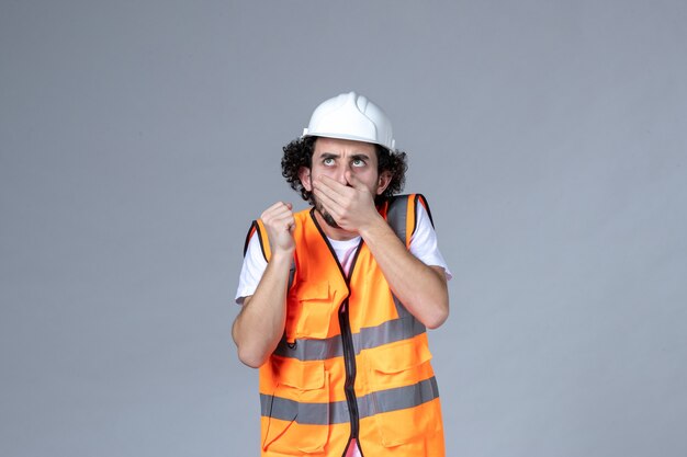 Vue de face du constructeur masculin pensant portant un gilet d'avertissement avec un casque de sécurité sur un mur gris vague