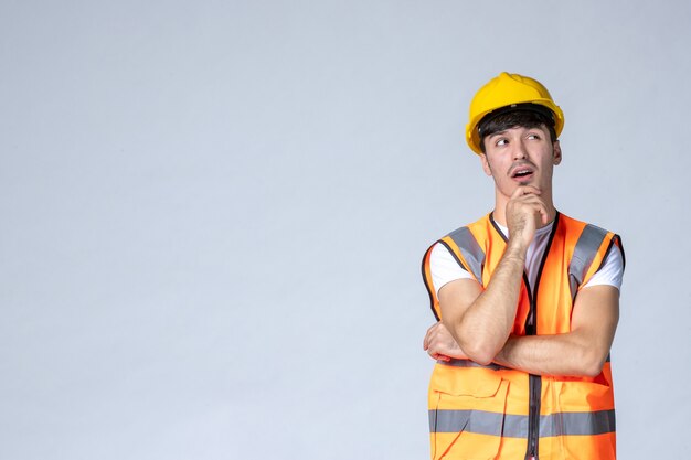 vue de face du constructeur masculin en uniforme et casque jaune pensant sur un mur blanc