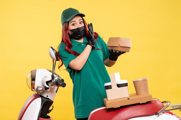 Vue de face du courrier féminin en masque avec livraison de café et de nourriture sur mur jaune